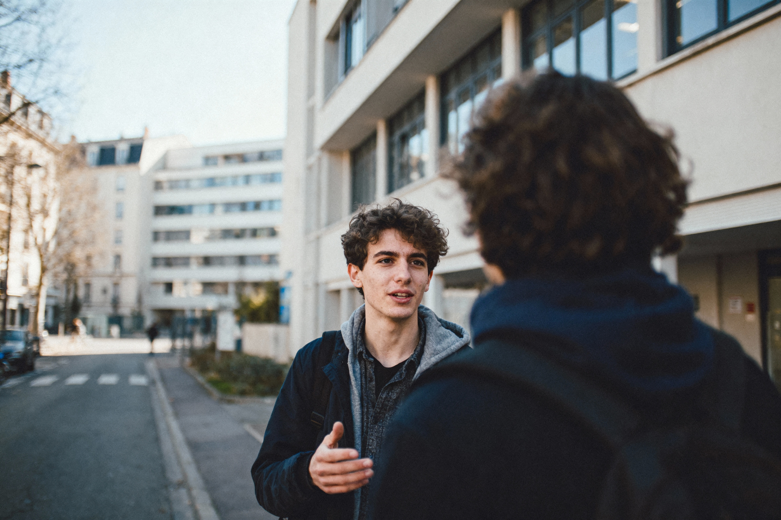 pexels-michal-pech-213601-757318 (1) Deux jeunes hommes discutent dans une rue urbaine, devant des bâtiments modernes, un jour ensoleillé. L'ambiance est détendue.