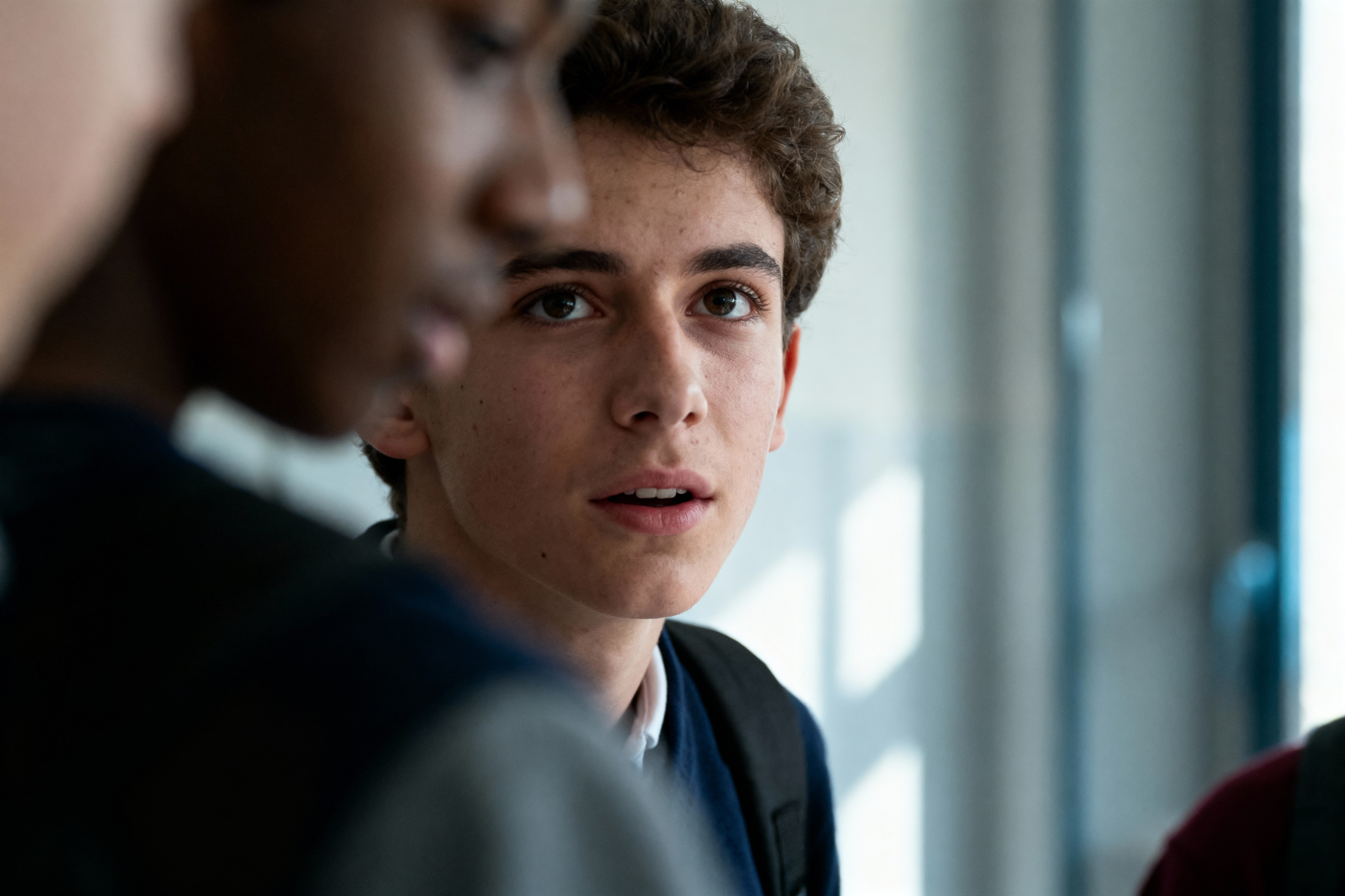 Un adolescent aux cheveux bouclés regarde intensément, entouré de camarades flous, dans un couloir lumineux.