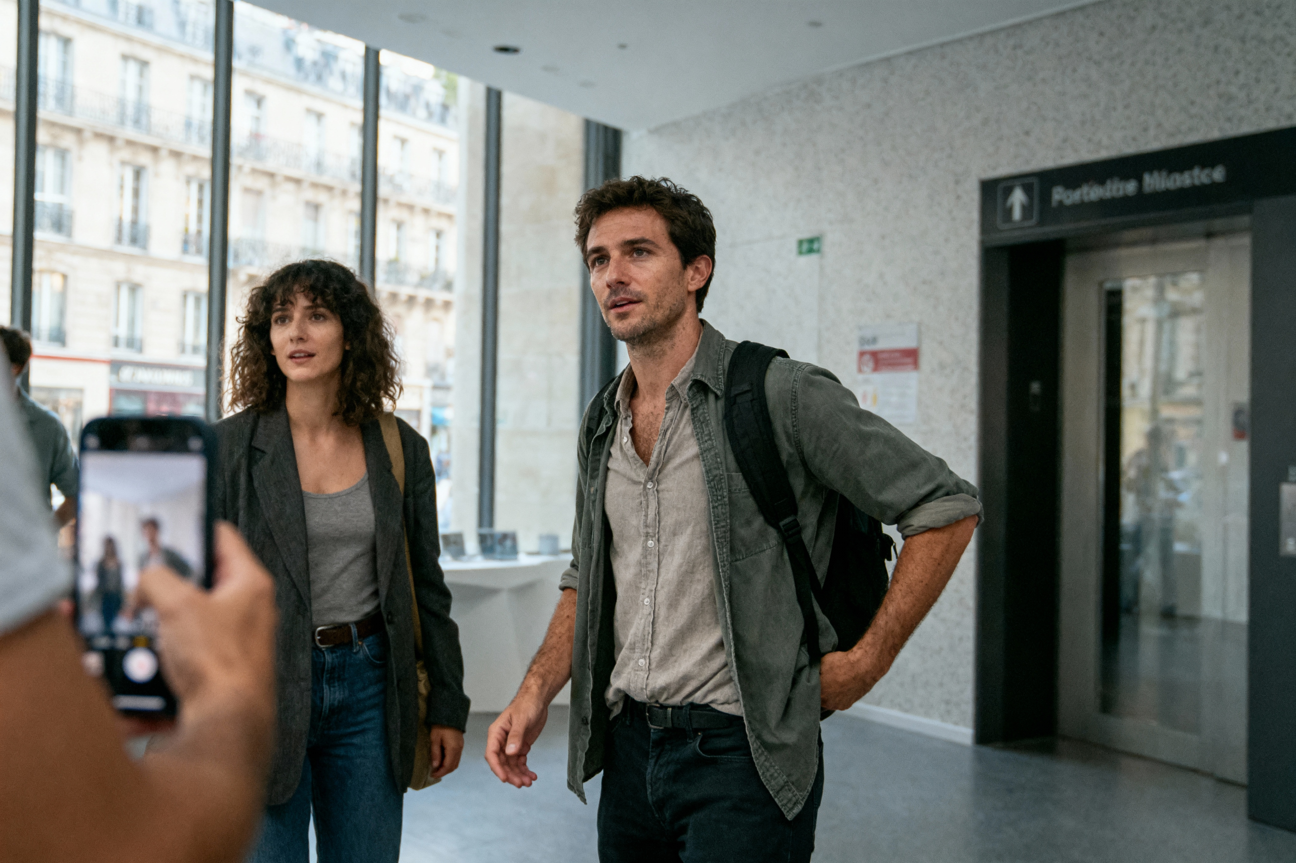 Deux personnes discutent dans un espace moderne avec des grandes fenêtres. Un homme et une femme, tous deux en tenue décontractée.