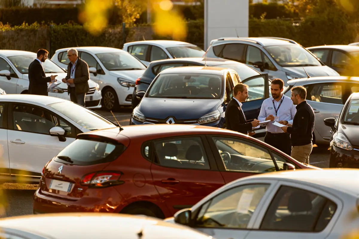 pexels-michal-pech-213601-757318 (1) Des personnes discutent dans un parking entouré de voitures, avec des documents à la main.