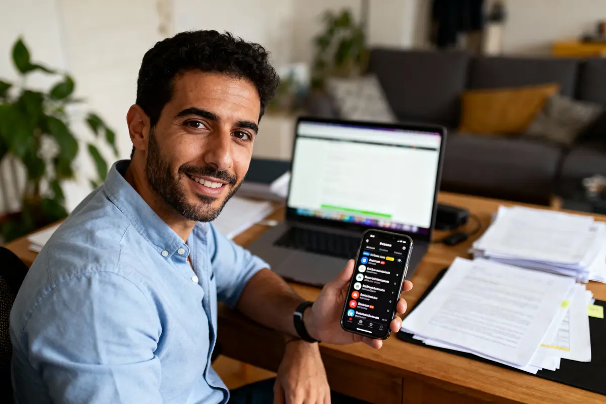 pexels-michal-pech-213601-757318 (1) Homme souriant tenant un smartphone, assis devant un ordinateur portable, bureau rempli de documents, intérieur de maison.