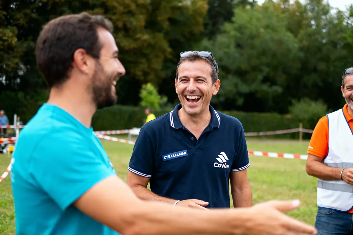 Trois hommes discutent et rient joyeusement à l'extérieur devant des arbres.