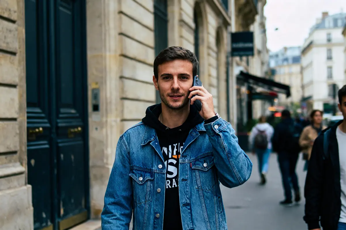 Jeune homme téléphone en marchant, portant une veste en jean, rue animée, devant bâtiments parisiens.