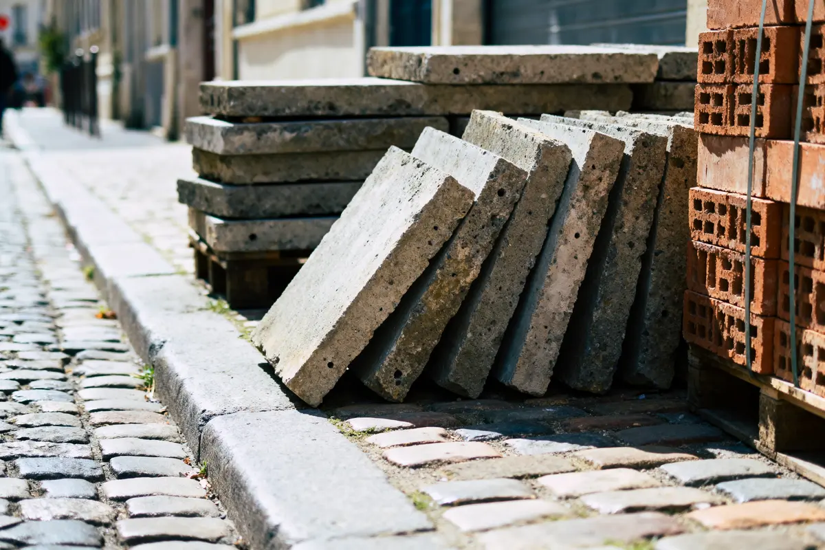 Dalles de béton et briques empilées sur un trottoir pavé, dans une rue urbaine ensoleillée.